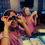 Two children participate in a hands-on STEM activity at a gaming center, one looking through small binocular-style lenses while the other assembles an electronic device, with gaming PCs and equipment visible in the background.