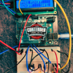 Close-up of a hands-on electronics project featuring a microcontroller board connected to a breadboard with multicolored wires and resistors, alongside a small LCD screen displaying text, representing coding and circuit-building in a STEM learning activity.